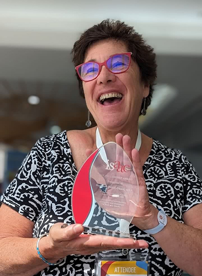 Dr. Fried-Oken holds up a clear glass award with red and white lettering 'isaac the fellowship award' wearing a black and white top, and a wide open smile that reaches her eyes behind red rimmed glasses that match the red accent. 